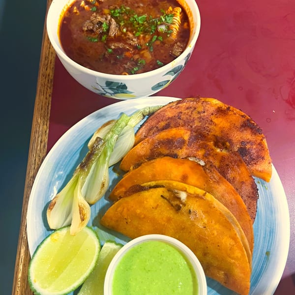 Birria tacos with consommé, grilled onions, lime, and green sauce on a red table.