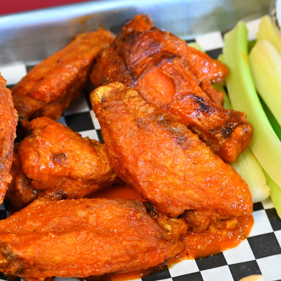 Pile of crispy buffalo chicken wings coated in orange sauce on a black-and-white checkered plate with celery sticks.